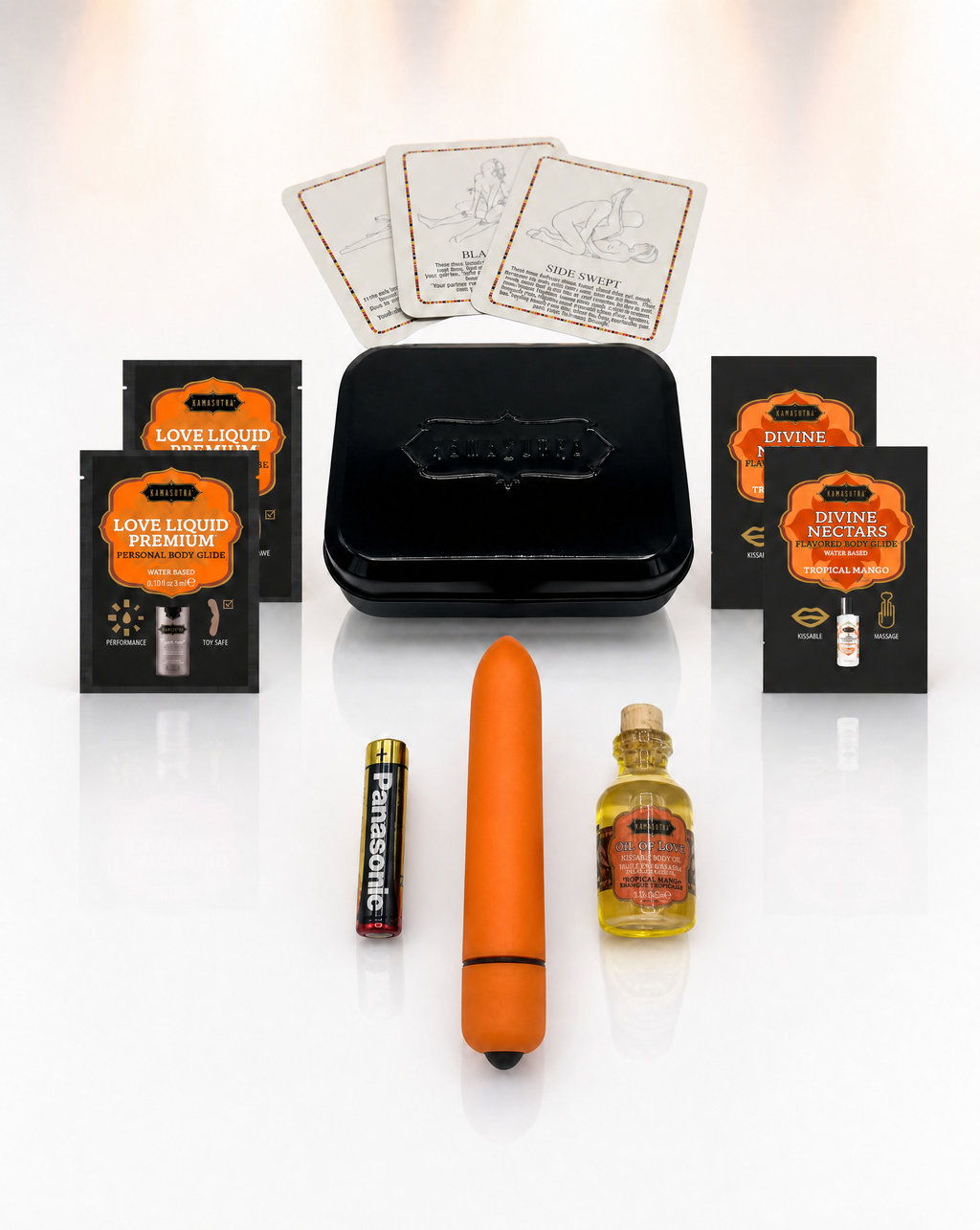 A photo showing the contents of the Kama Sutra weekender vibe kit in Tropical Mango. There is 3 position cards, 2 Love Liqud packets, two divine nectar Tropical Mango packets, an orange bullet vibe with battery and a mini glass Oil of Love outside a black tin.