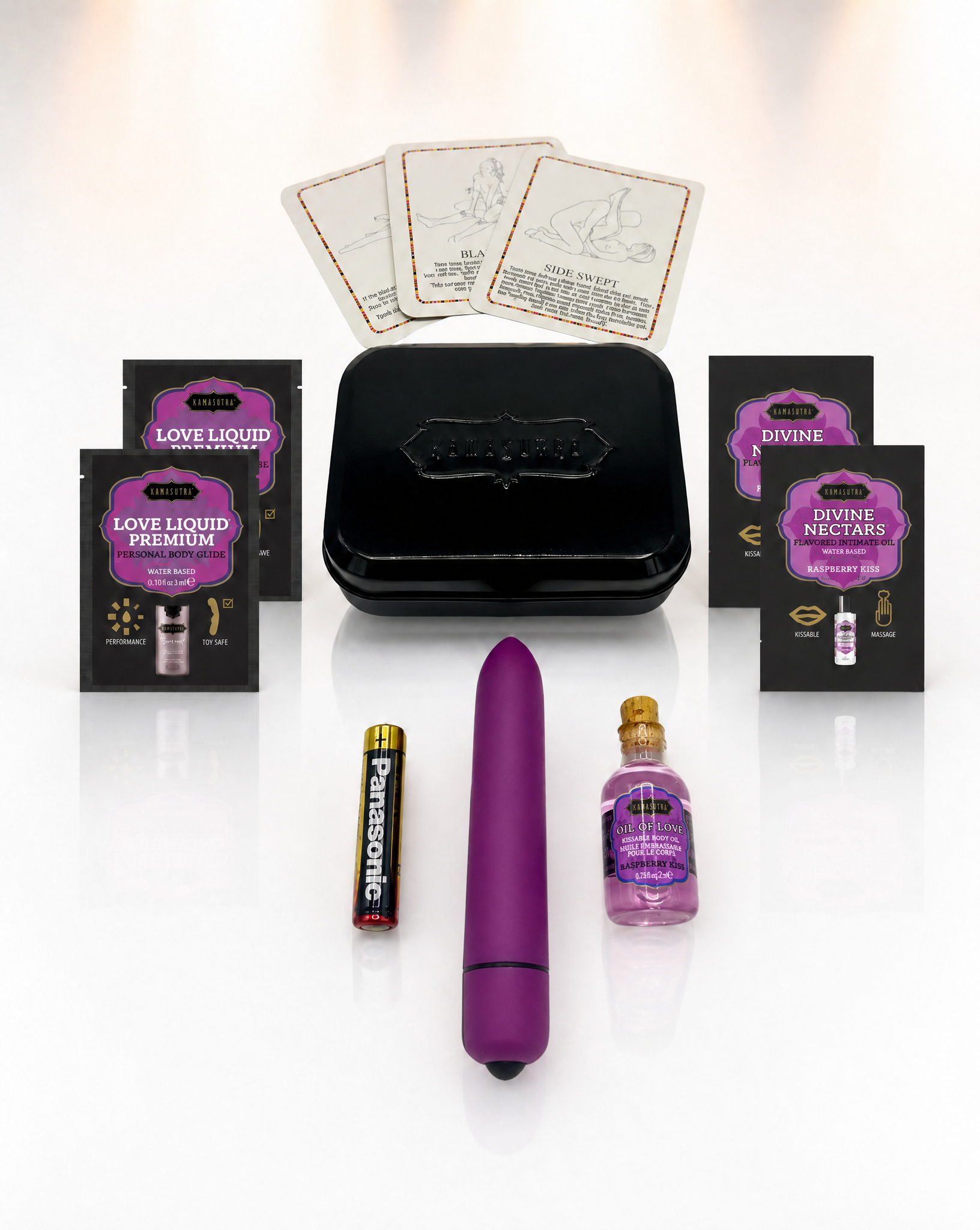 A photo showing the contents of the Kama Sutra weekender vibe kit in Raspberry Kiss. There is 3 position cards, 2 Love Liqud packets, two Divine Nectar Raspberry Kiss packets, a purple bullet vibe with battery and a mini glass Oil of Love outside a black tin.
