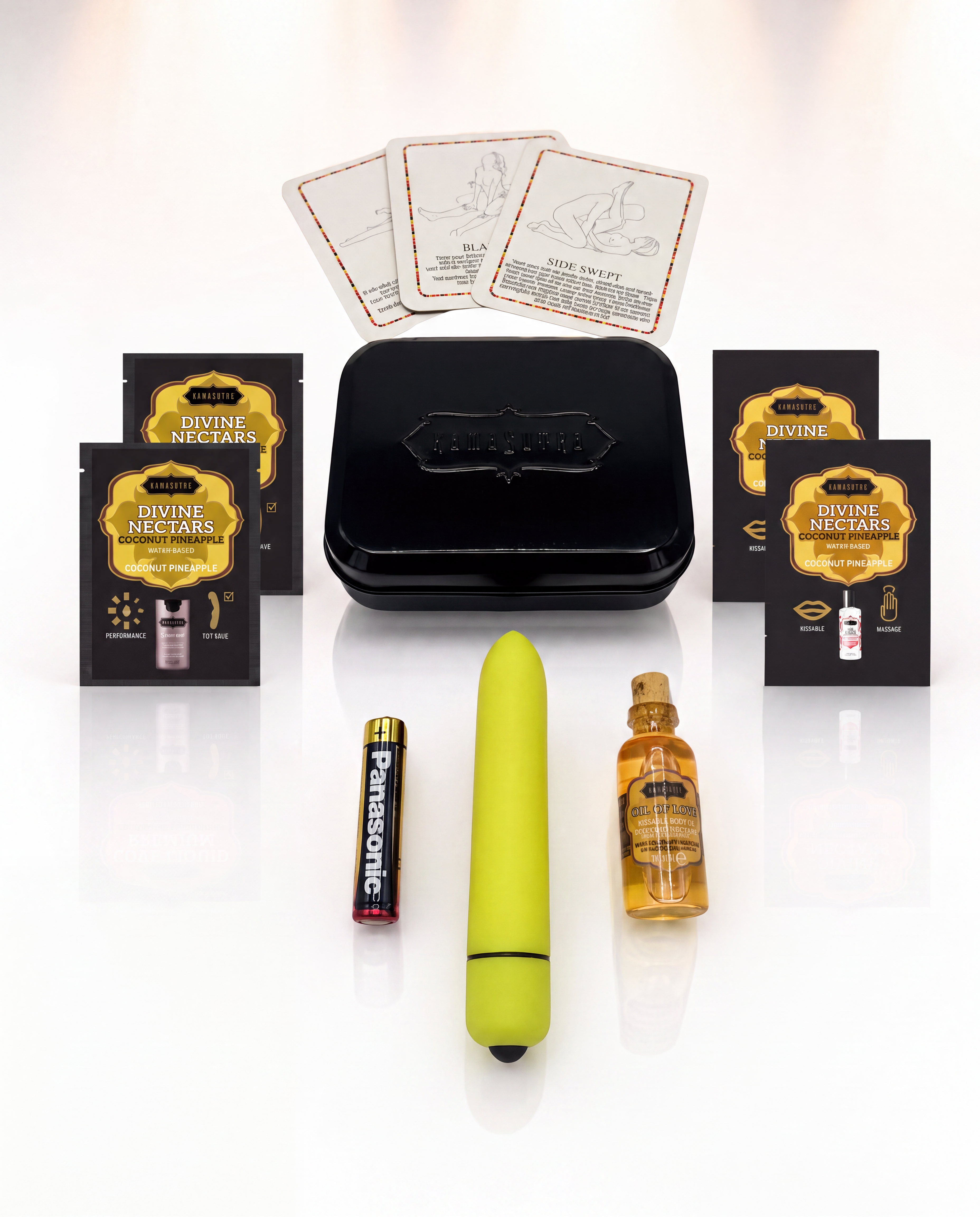 A photo showing the contents of the Kama Sutra weekender vibe kit in Coconut Pineapple. There is 3 position cards, 2 Love Liqud packets, two Divine Nectar Coconut Pineapple packets, a yellow bullet vibe with battery and a mini glass Oil of Love outside a black tin.